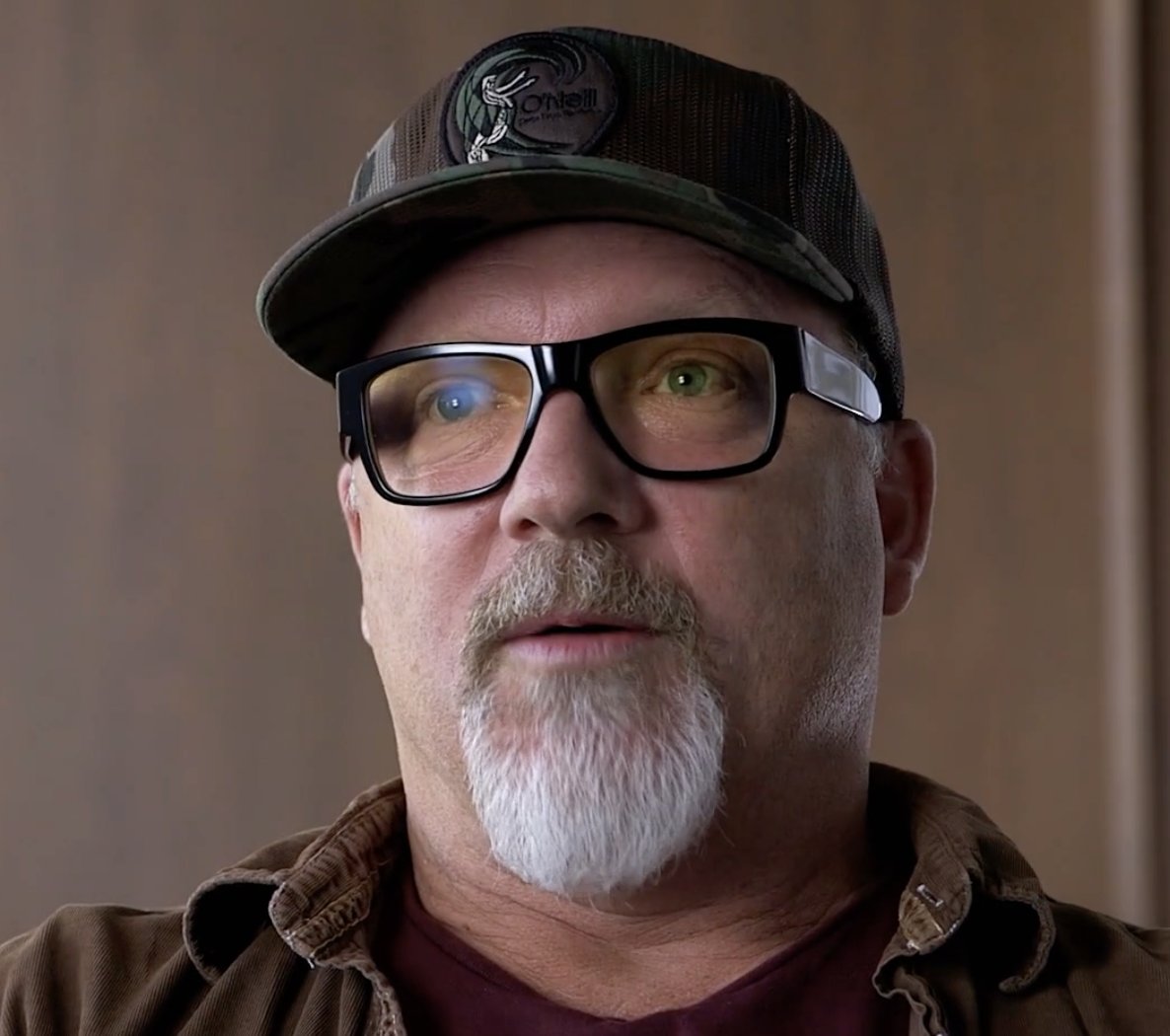 John Boiler in a camo cap and black-framed glasses, mid-interview, against a wood-panelled wall.