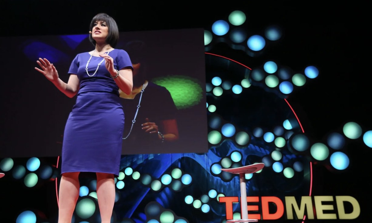 Lisa Shufro speaking on the TEDMED stage in a blue dress, lit by a wall of coloured lights.