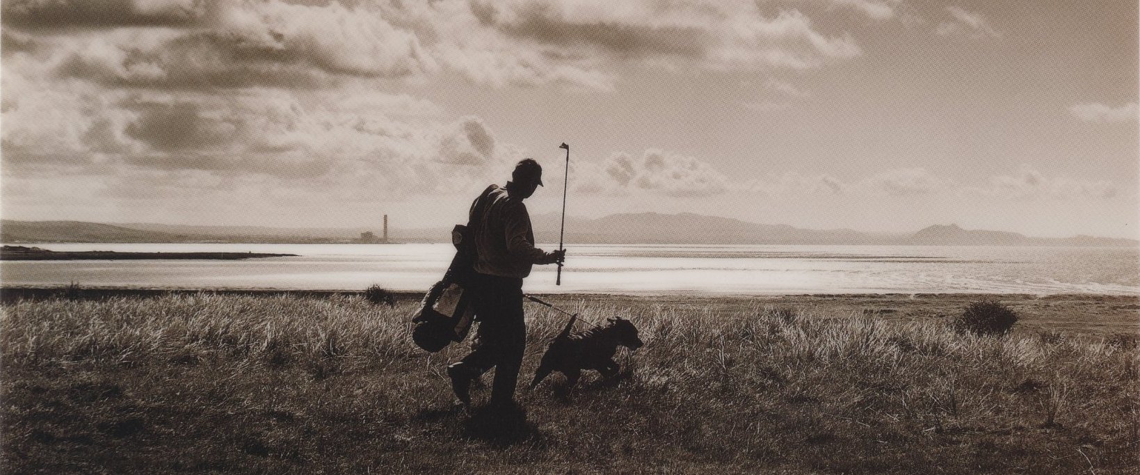 Sepia photograph from the Golf is an Invitation campaign: a lone golfer carrying his bag walks a windswept links beside the sea, a dog trotting ahead.