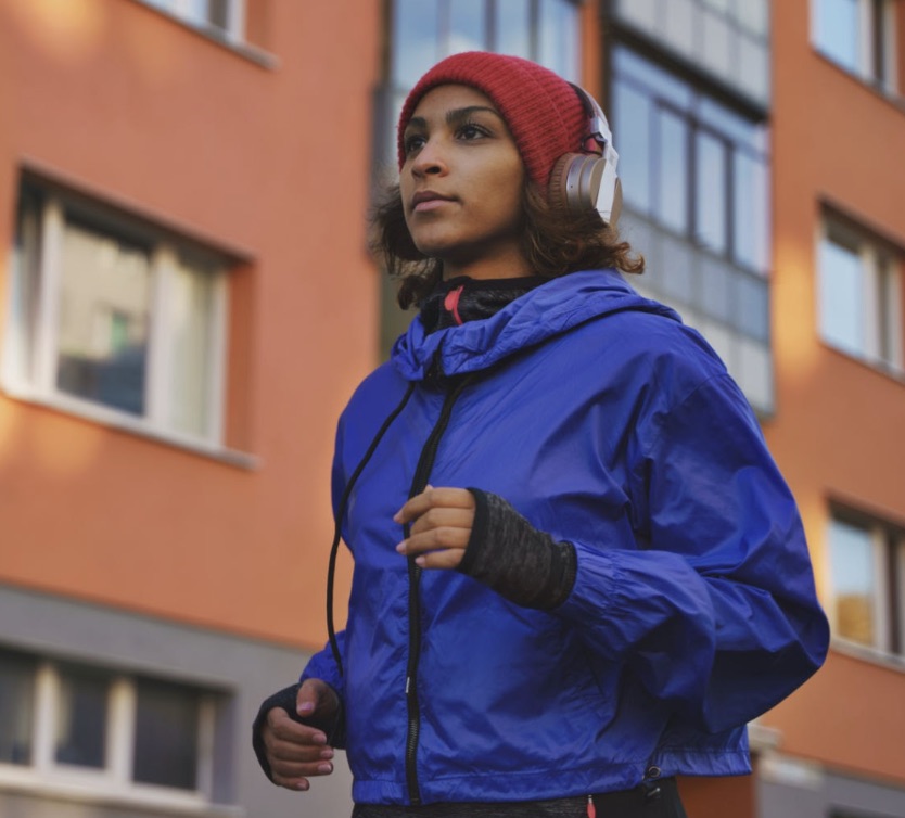 A runner in a blue jacket and red beanie, headphones on, mid-stride.